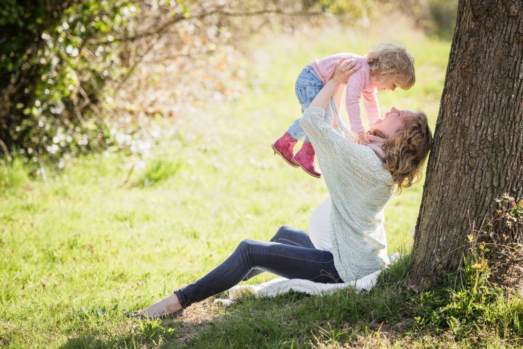A pregnant woman sitting against a tree, joyfully lifting her young daughter in the air in a sunlit grassy area.